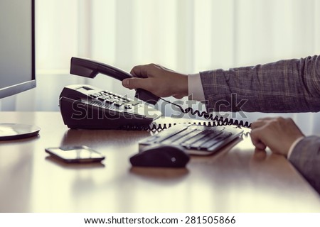 Businessman pick up or hangs up the phone in the office, keyboard, mouse, mobile, and monitor detail in the background, with vintage color tone effect.
