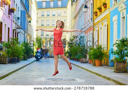 Beautiful young woman in red polka dot dress jumping on a Parisian street with colorful bright houses