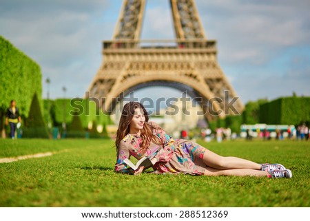 Beautiful young woman walking in Paris lying on the grass near the Eiffel tower on a nice spring or summer day and reading a book. Student or schoolgirl doing her homework or preparing to exams