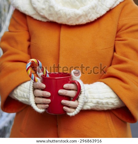 Woman hands in woolen mittens holding a cozy mug with hot cocoa, tea or coffee and a Christmas candy cane