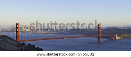 A panoramic image of Golden Gate Bridge lit by the setting sun with the San Francisco skyline in the background.