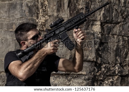 View of a menacing man pointing a machine gun in a black shirt and dark shades on a stone quarry.