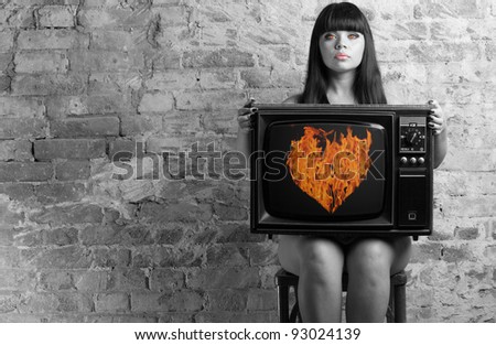 The woman in the image of the devil keeps an old television with the image of a fiery heart on the screen.