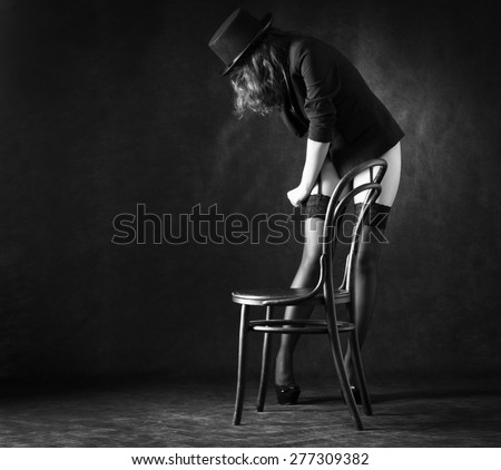 Sexy woman in black stockings and hat is dancing around a chair on a dark background.