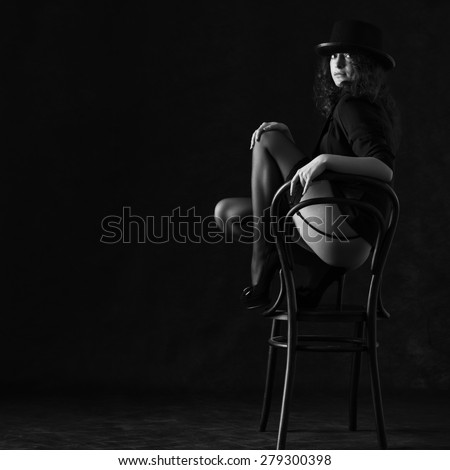Sexy woman in black stockings and hat is dancing around a chair on a dark background.