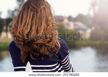 Young woman with long curly hair standing with her back looking at the water scenery.