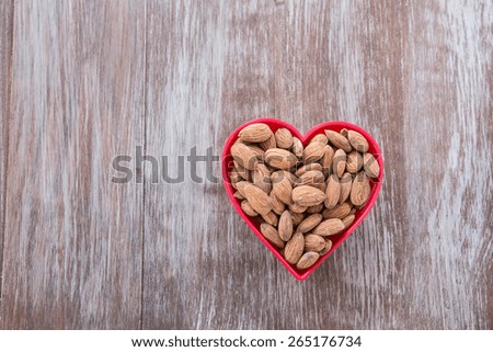 A heap of almonds in a red heart shaped bowl on wood background from above
