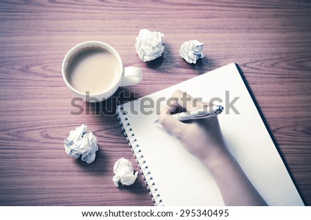 woman hand writing with pen on notebook.there are crumpled paper and coffee cup on wood table background vintage style