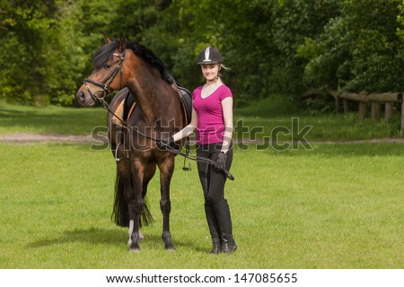 Girl stands besides her brown pony (New-Forest-Pony) on a meadow