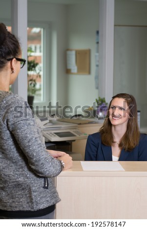 Smiling clerk (caucasian) is sitting behind a counter and is looking to a customer (latin) in front of the counter. Woman in the background is in focus, woman in the foreground is blurred.