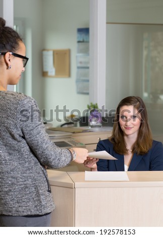 Clerk (caucasian) is handing over a document to a client (latin) at a reception counter in the office. Woman in the background is in focus, woman in the foreground is blurred.