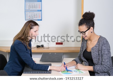 Two business women are in a meeting and are sitting at a table in the office. Both women are pointing to a document.