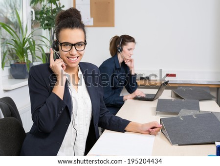 A young business woman with headset is sitting in front of her files. Another woman with headset is sitting in the background (blurred).