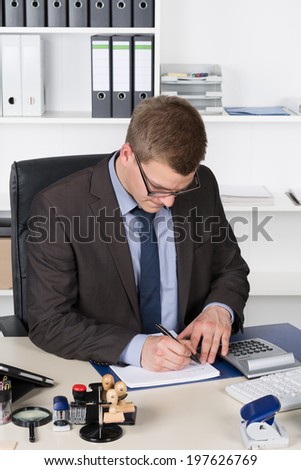 Young businessman is writing into a file while sitting at the desk in the office. The man is looking to the file.