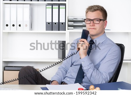 Young businessman with glasses is holding a telephone receiver while sitting relaxed at the desk in the office.