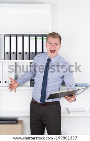 Young furious businessman is holding an opened file while standing in front of a shelf in the office. The man is looking to the camera.