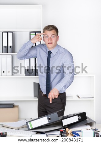 Young furious businessman is standing in front of many files on his desk in the office. He is pointing with one finger to his head and with the other to the files in front of him.
