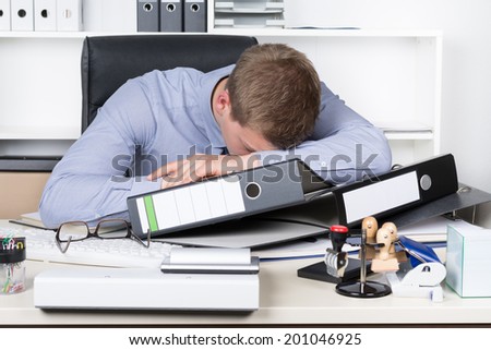 Young exhausted businessman is lying on his desk in the office in front of many files. A shelf is in the background. The man is looking down.