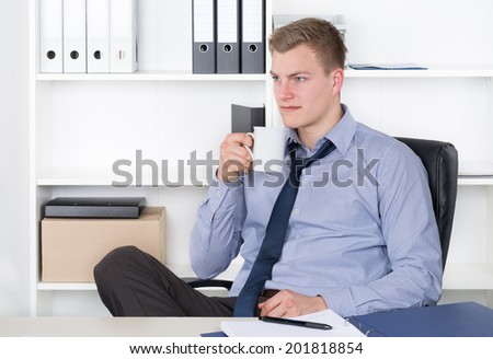 Young relaxed businessman is doing a coffee break while sitting at the desk in the office. A shelf is in the background. The man is looking sideways.
