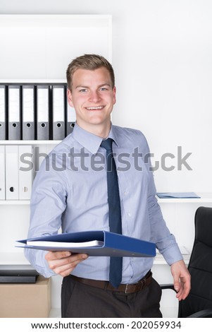 Young smiling businessman is handing over a file while standing in the office. A shelf is in the background. The man is looking to the camera.