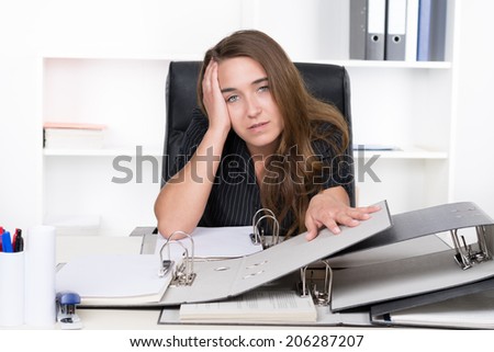 A young frustrated businesswoman is sitting in front of a pile of files at the desk in the office. A shelf is standing in the background. The woman is looking to the camera.