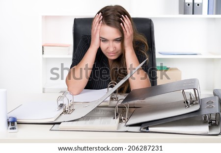 A young frustrated businesswoman is sitting in front of a pile of files at the desk in the office. A shelf is standing in the background. The woman is looking down.