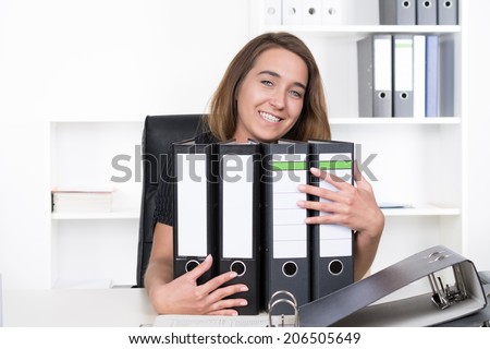 A young smiling businesswoman is sitting in front of several files at the desk in the office. A shelf is standing in the background. The woman is looking to the camera.