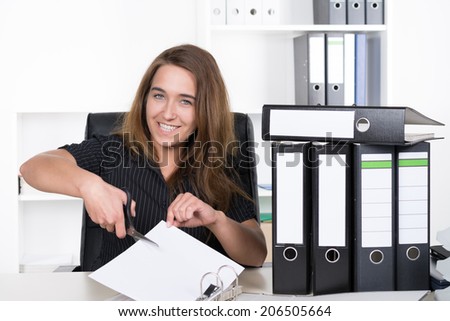 A young smiling businesswoman is cutting a sheet of paper with a pair of scissors while sitting besides several files at the desk in the office. A shelf is standing in the background.