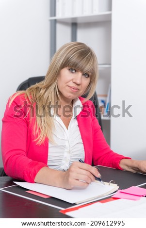 Woman is writing into a file while sitting at the desk in the office. The woman is looking to the camera.