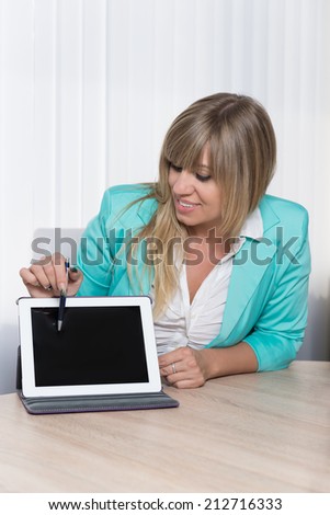 Smiling businesswoman is pointing with a pen to a tablet while sitting at a table in the office. The woman is looking to the tablet.