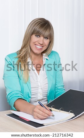 Smiling businesswoman is writing into a file while sitting at a table in the office. The woman is looking to the camera.