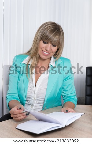 A businesswoman is looking into an appointment calendar which is lying in front of her while she is sitting at a table in the office.
