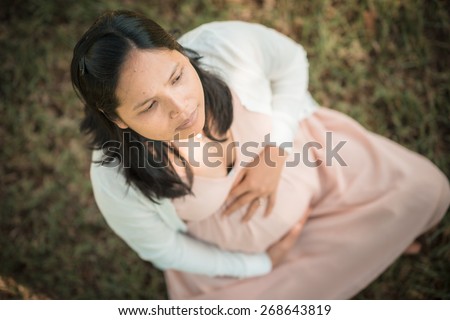Asian woman 9 months pregnant sitting outside in the grass holding her belly looking serious. Shallow focus on eyes to mouth. Looking off camera. Muted tones, soft faded filter effects.