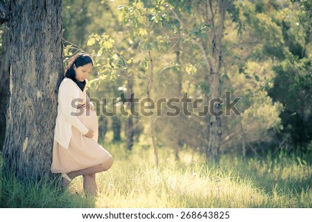 Asian woman 9 months pregnant standing outside among trees in long grass. Tranquil and natural environment. Muted tones, soft faded filtered effects.