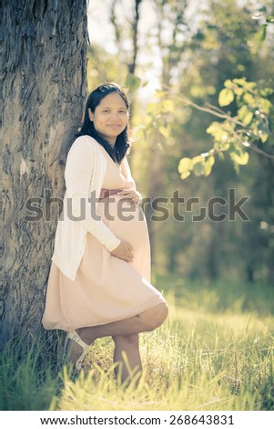 Asian woman 9 months pregnant standing outside among trees in long grass. Tranquil and natural environment. Muted tones, soft faded filtered effects.