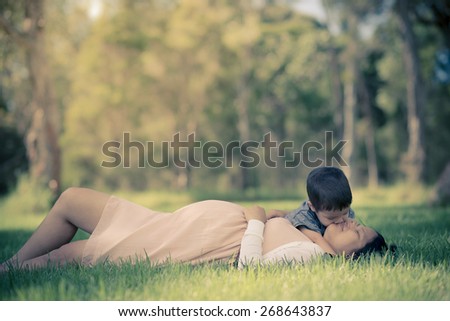 Asian woman 9 months pregnant laying outside in the grass with her 2 year old son kissing her. Muted tones, faded filtered effects