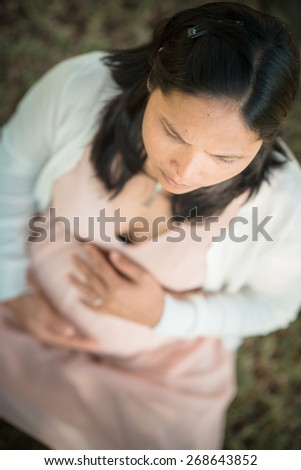 Asian woman 9 months pregnant sitting outside in the grass holding her belly looking serious. Shallow focus on eye lashes. Looking off camera. Muted tones, soft faded filter effects.