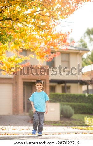 Cute 5 year old mixed race Asian Caucasian boy walks across the street of his suburban neighborhood with beautiful Autumn (Fall) trees in the background