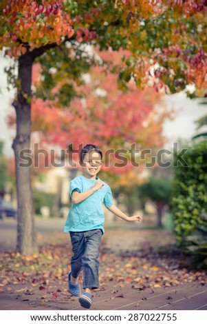 Cute 5 year old mixed race Asian Caucasian boy runs down the footpath (sidewalk) of his suburban neighborhood with beautiful Autumn (Fall) trees lining the street
