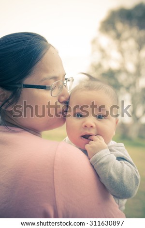 Cute 4 month old mixed race Asian Caucasian Thai baby snuggles with his Thai Asian mother outdoors in the late afternoon sun in Australia. Filtered color shift, sun flare, and soft focus effects