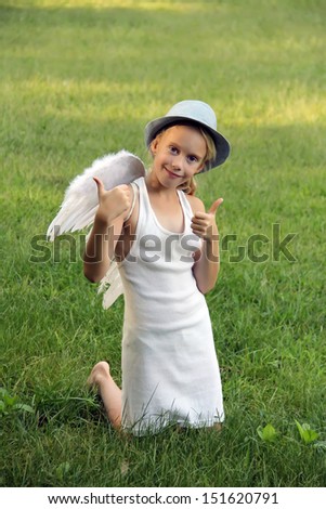 Cute little girl with angel wings shows thumbs up sitting on the green lawn