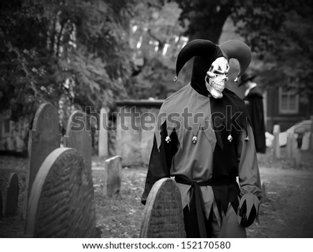 Scary Joker with skeleton face looking at the headstone on the graveyard
