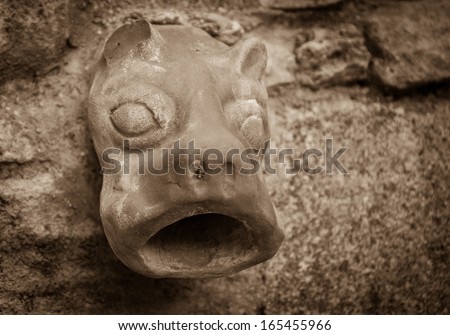 Old ceramic downspout in shape of beast head against stone wall. Closeup. Sepia.