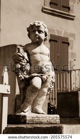 Statue of Bacchus (Dionysus) with grapes in his hands. Garden sculpture and old stone house with maroon wooden shutters at background. Chateauneuf du Pape, Provence, France. Aged photo. Sepia.