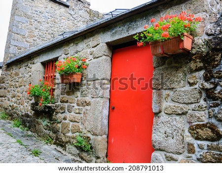 Old stone house with red door, red painted grilles on the windows and hanging boxes with red geranium flowers. Medieval town Moncontour. Brittany, France