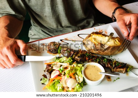 Delicious beef and duck kebab with fresh green salad, baked potato and French pepper cream sauce. Young man's hands with fork and knife.
