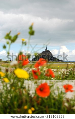 Mont Saint Michel UNESCO heritage site. Poppy flowers field under cloudy sky. Tourists going towards Mont Saint Michel. Normandy, France. Selective focus on the Mont Saint Michel.