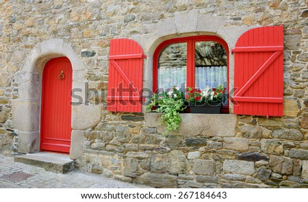 Old stone house with red wooden shutters and red door. Boxes with red and white flowers on the window. Brittany, France