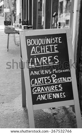 Signboard outside old books store in France. Text in French meaning: Secondhand bookseller buying old and modern books, postcards, ancient prints. Aged photo. Black and white.