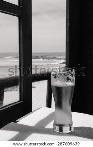 Cold beer on cafe terrace with the view on the ocean beach through the opened window. Algarve, Portugal. A game of light an shadow. Aged photo. Black and white.